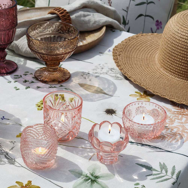 Vier Chic Antique Teelichthalter Glas Rosa Vintage Ø8 cm mit brennenden Teelichtern auf einer geblümten Tischdecke, umgeben von Glaswaren, einem Strohhut und einem gefalteten Tuch.