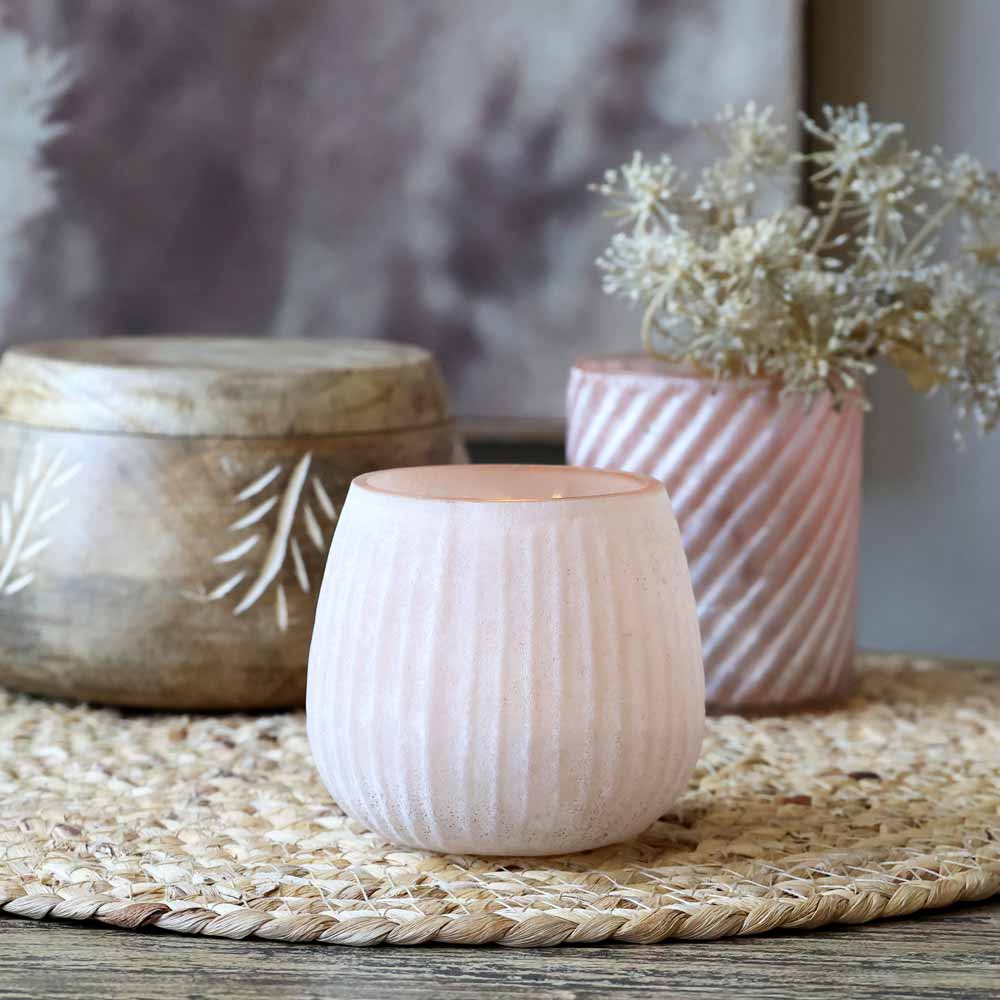Ein kleiner Chic Antique-Teelichthalter mit Rillen rosa steht auf einer geflochtenen Matte, begleitet von einem größeren Holzbehälter mit Farnmuster und einer rosa Vase mit getrockneten Blumen im Hintergrund.