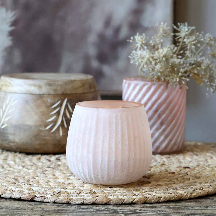 Ein kleiner Chic Antique-Teelichthalter mit Rillen rosa steht auf einer geflochtenen Matte, begleitet von einem größeren Holzbehälter mit Farnmuster und einer rosa Vase mit getrockneten Blumen im Hintergrund.
