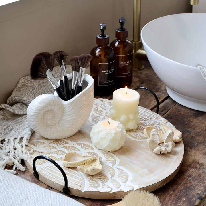 Auf einem Holztablett stehen die Chic Antique Vase Muschel (muschelförmig), Make-up-Pinsel, zwei brennende Kerzen, Dekoartikel, braune Flaschen und ein Handtuch neben einem weißen Waschbecken.