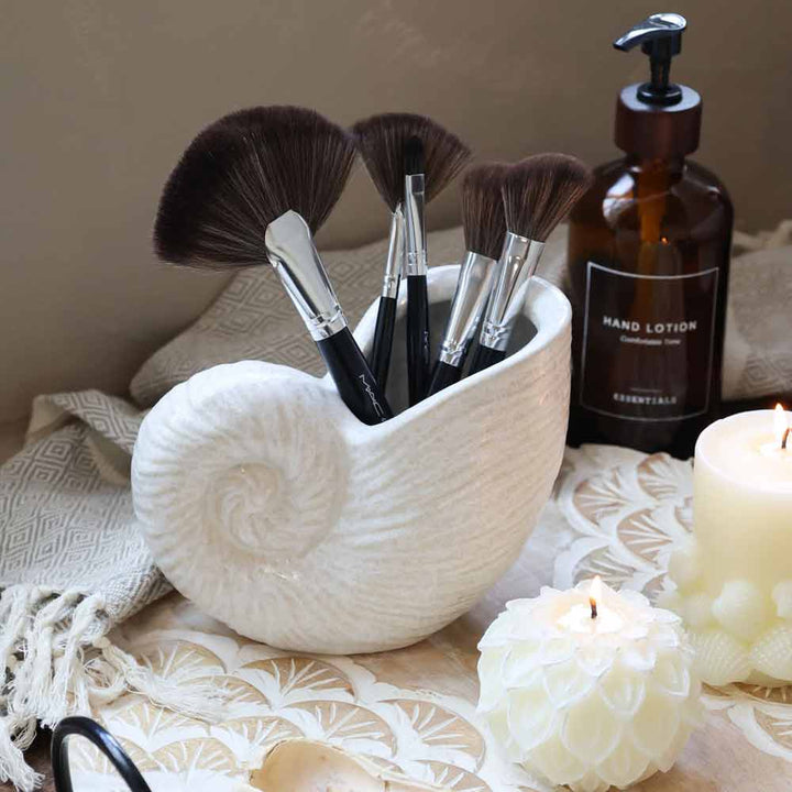 Fünf Make-up-Pinsel, ausgestellt in der Chic Antique – Vase Muschel, einem weißen muschelförmigen Halter, auf einem Tisch mit einer Kerze, einer Handlotionflasche und einem strukturierten Tuch.