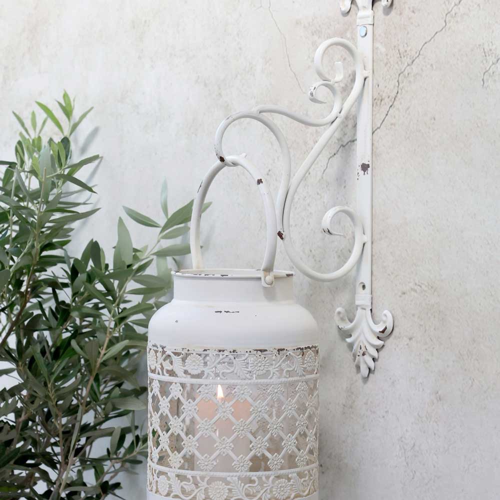 Eine weiße Laterne im Vintage-Stil hängt an einer Chic Antique-Wandhalterung für Laternen an einer strukturierten Wand, in der Nähe von grünem Laub.