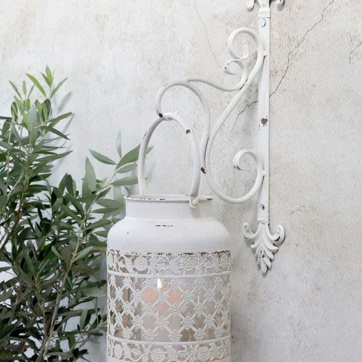 Eine weiße Laterne im Vintage-Stil hängt an einer Chic Antique-Wandhalterung für Laternen an einer strukturierten Wand, in der Nähe von grünem Laub.