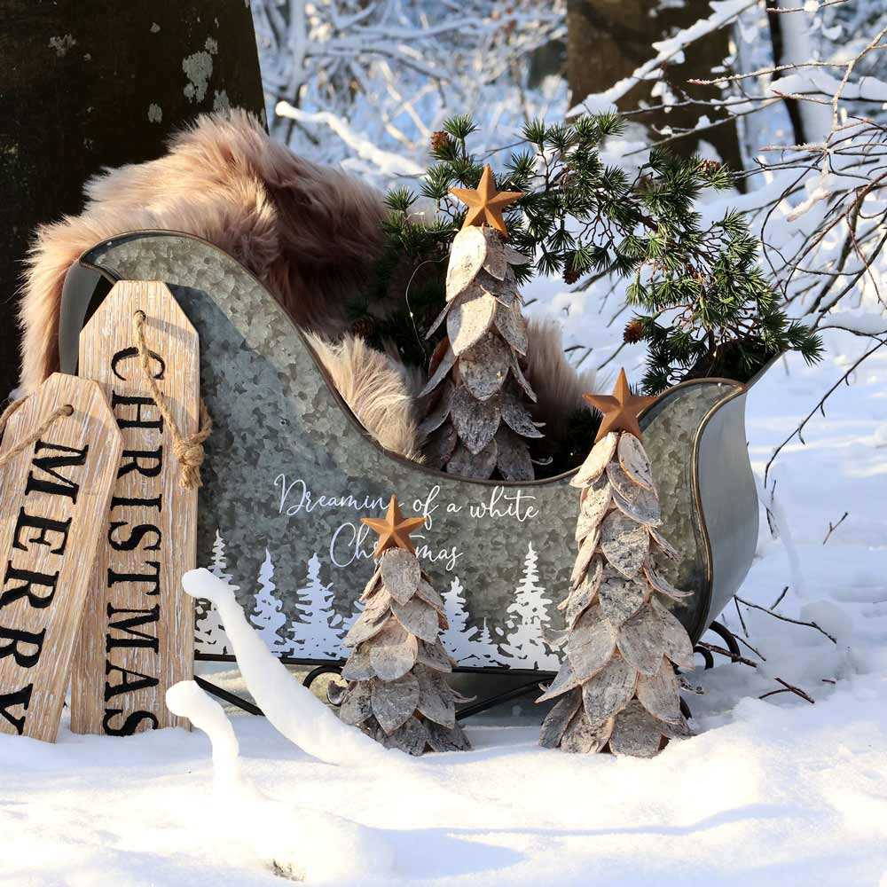 Chic Antique – Weihnachtsbaum mit Stern, arrangiert mit Tannenzweigen, hölzernen Weihnachtsbäumen und dekorativen Schildern in einer verschneiten Waldkulisse.