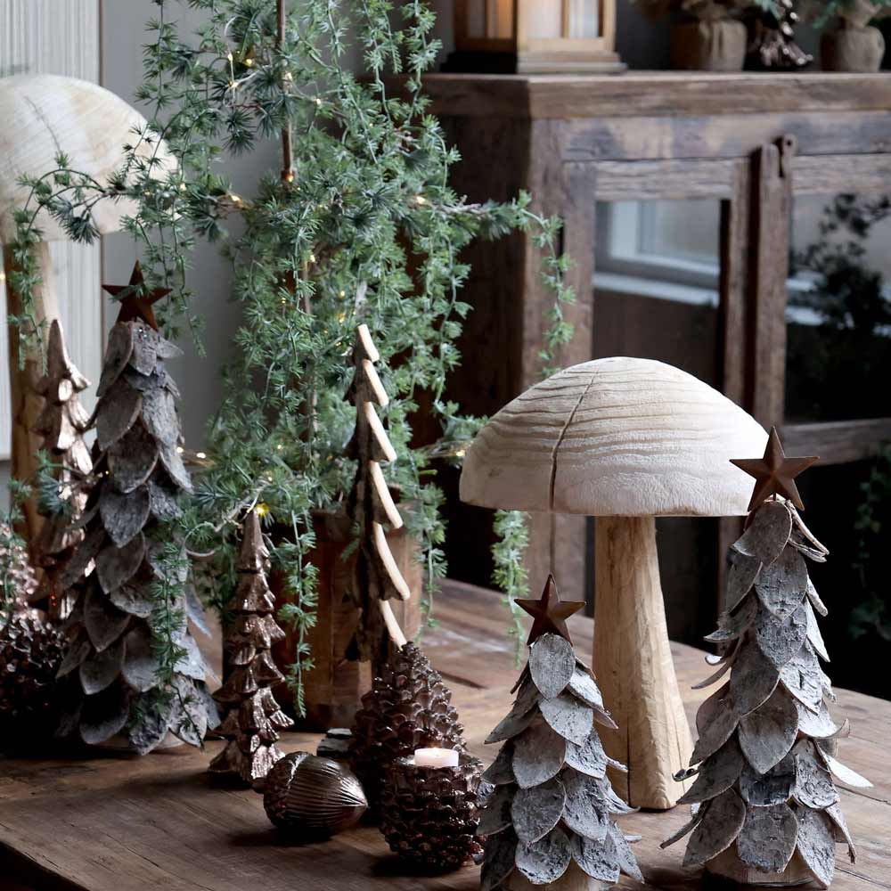 Eine Chic Antique-Ausstellung mit einem Weihnachtsbaum mit Stern, begleitet von kleinen Holzbäumen, Tannenzapfen, einem geschnitzten Pilz und immergrünen Zweigen auf einem Holztisch.