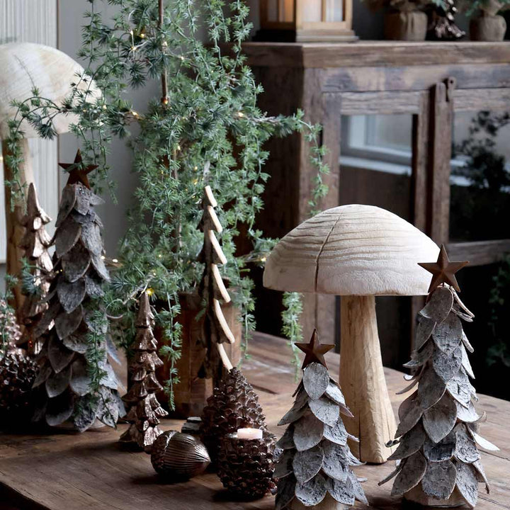 Eine Chic Antique-Ausstellung mit einem Weihnachtsbaum mit Stern, begleitet von kleinen Holzbäumen, Tannenzapfen, einem geschnitzten Pilz und immergrünen Zweigen auf einem Holztisch.