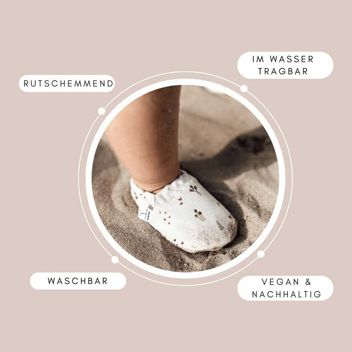 Ein Fuß mit Cosy Roots – Lauflernschuhen. Regenbogen stehen auf Sand, umgeben von Etiketten mit hervorhebenden Merkmalen: „rutschhemmend“, „im wasser tragbar“, „waschbar“, „vegan & nachhaltig“.