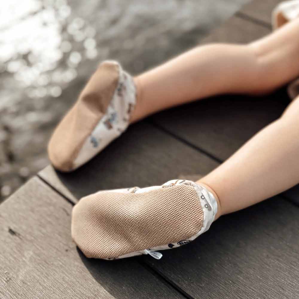 Eine Person sitzt auf einem Holzdeck am Wasser und trägt Cozy Roots - Lauflernschuhe Little Driver mit Blumenmuster.