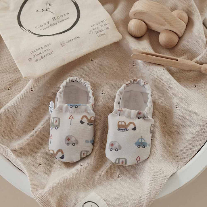 Ein Paar Cosy Roots - Lauflernschuhe Little Driver mit Fahrzeugdruck auf beigem Textiluntergrund, dazu Holzspielzeug und ein dekoratives rundes Schild.