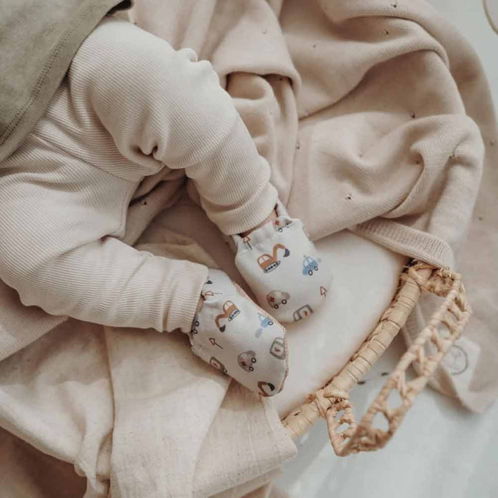 Baby trägt Cozy Roots - Lauflernschuhe Little Driver mit Tiermuster sitzt in einem geflochtenen Korb.