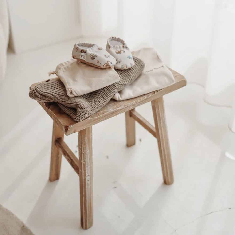 Ein Paar Cozy Roots - Lauflernschuhe Little Driver auf einer gefalteten Decke, platziert auf einem kleinen Holzhocker in einem hell beleuchteten Raum.