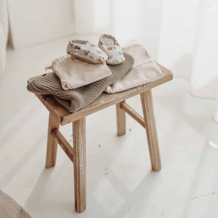 Ein Paar Cozy Roots - Lauflernschuhe Little Driver auf einer gefalteten Decke, platziert auf einem kleinen Holzhocker in einem hell beleuchteten Raum.