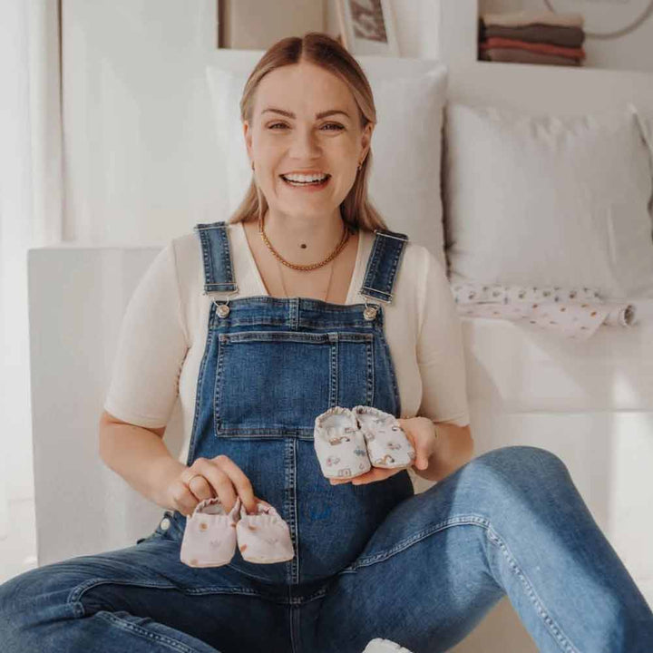 Eine lächelnde Frau sitzt auf einem Bett, hält Cozy Roots - Lauflernschuhe Little Driver in der Hand und trägt einen Jeansoverall.