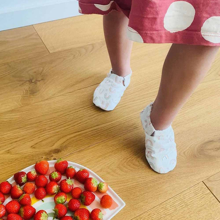 Ein Kleinkind steht auf einem Holzboden und trägt Cosy Roots-Lauflernschuhe mit Regenbogen und Wolkenmuster, neben einem Teller mit Erdbeeren.