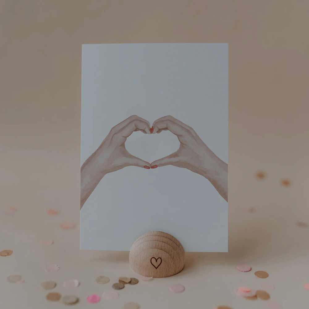 Die Eulenschnitt - Aquarellkarte Heart Hands zeigt zwei Hände, die ein Herz bilden, aufrecht in einem Holzhalter mit Herzsymbol präsentiert, umgeben von Konfetti auf einem neutralen Hintergrund.
