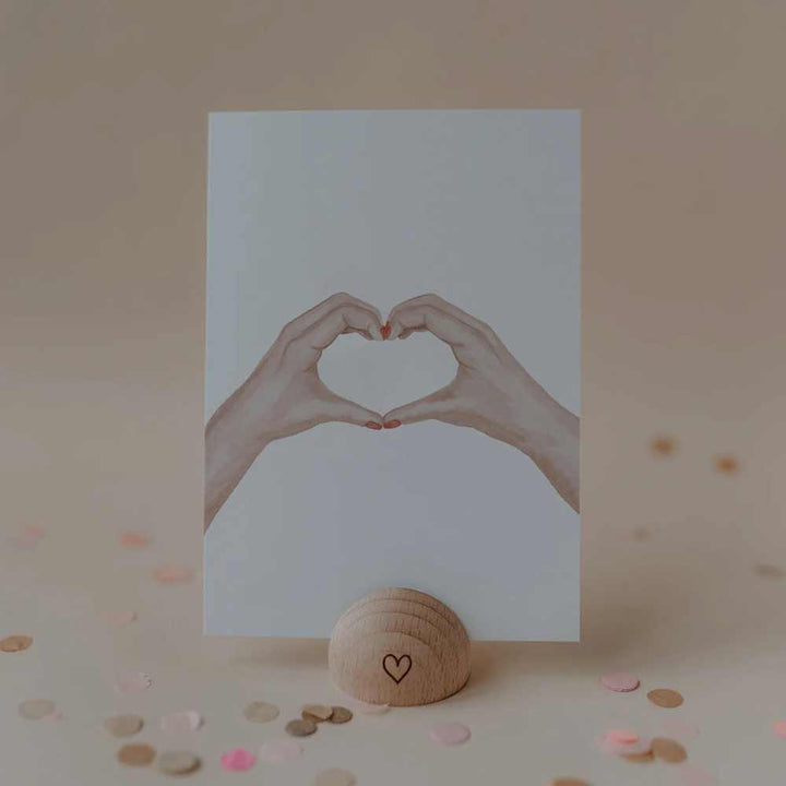 Die Eulenschnitt - Aquarellkarte Heart Hands zeigt zwei Hände, die ein Herz bilden, aufrecht in einem Holzhalter mit Herzsymbol präsentiert, umgeben von Konfetti auf einem neutralen Hintergrund.