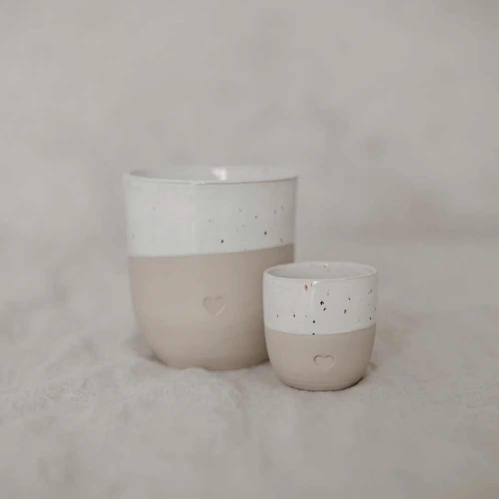 Der Eulenschnitt - Espresso Becher Herz besteht aus zwei Keramiktassen, einer großen und einer kleinen, in Weiß und Beige mit gesprenkeltem Muster und Herzdesign, dargestellt auf einer leicht strukturierten Oberfläche.