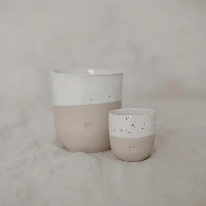 Der Eulenschnitt - Espresso Becher Herz besteht aus zwei Keramiktassen, einer großen und einer kleinen, in Weiß und Beige mit gesprenkeltem Muster und Herzdesign, dargestellt auf einer leicht strukturierten Oberfläche.