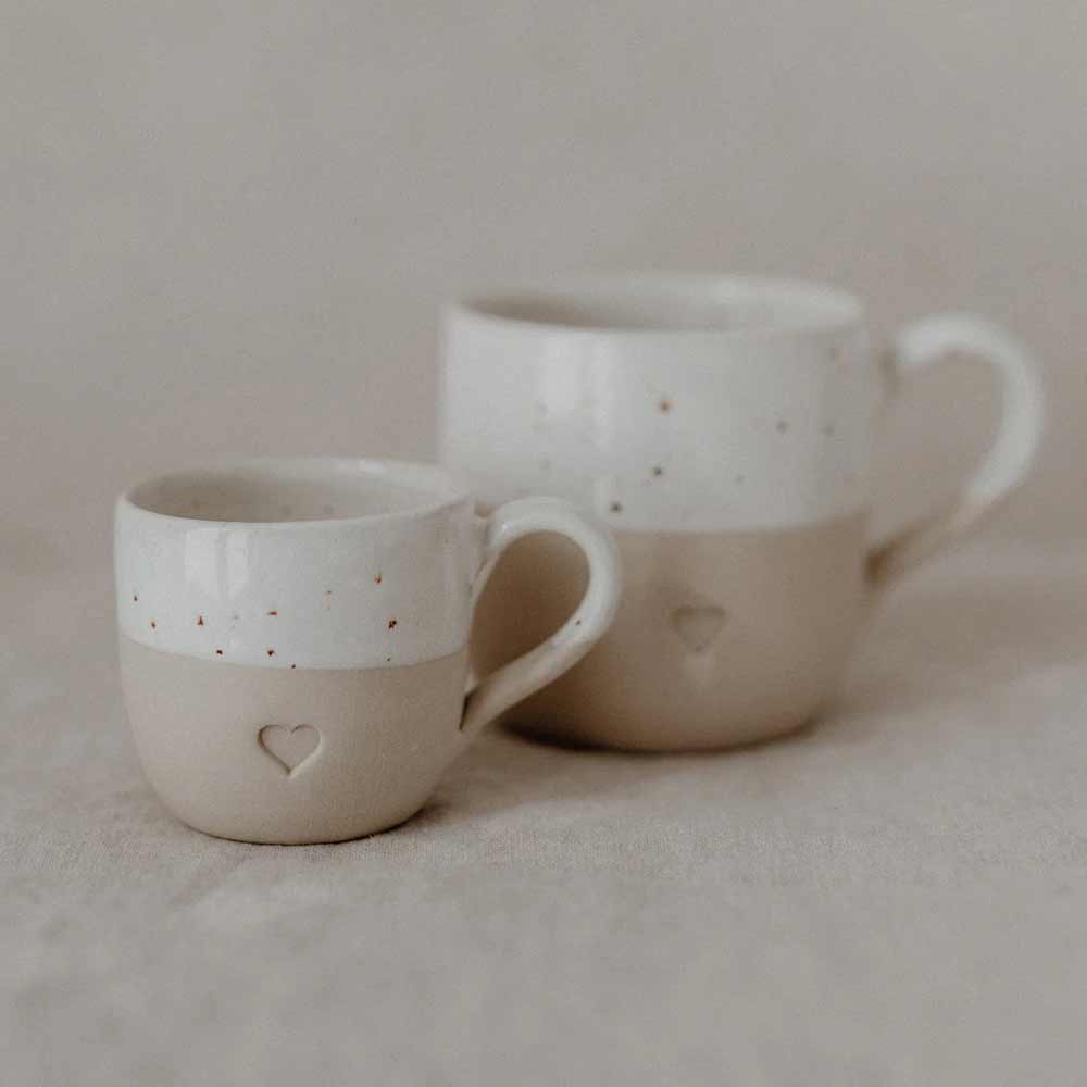 Zwei Eulenschnitt Espresso Tasse Herz, eine kleine und eine große, mit beige-weißer Glasur und geprägtem Herz, stehen auf einer neutralen Stoffoberfläche.
