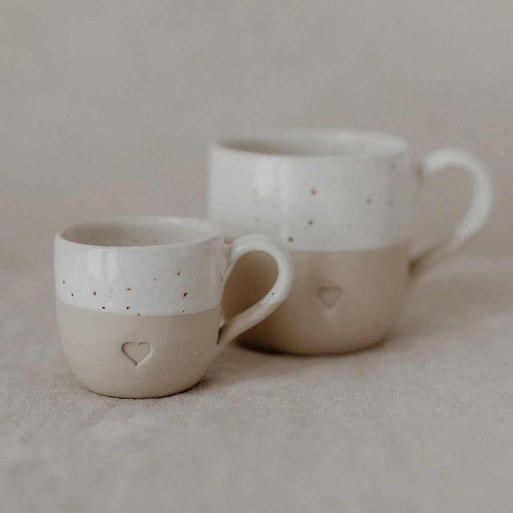 Zwei Eulenschnitt Espresso Tasse Herz, eine kleine und eine große, mit beige-weißer Glasur und geprägtem Herz, stehen auf einer neutralen Stoffoberfläche.