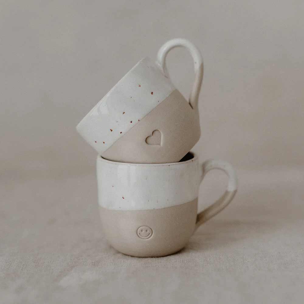 Zwei Eulenschnitt - Espresso Tasse Herz Keramikbecher sind gestapelt, einer mit einem Herz und der andere mit einem Smiley, vor einem neutralen Hintergrund.