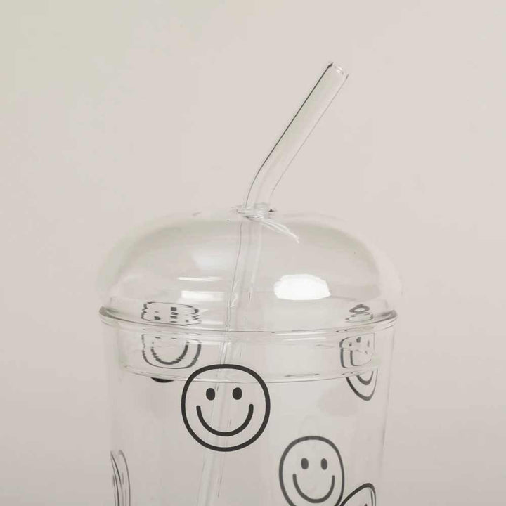 Ein durchsichtiger Plastikbecher mit gewölbtem Deckel (Eulenschnitt – Glasdeckel für Trinkglas), mit schwarzen Smiley-Motiven und einem gebogenen, durchsichtigen Strohhalm.