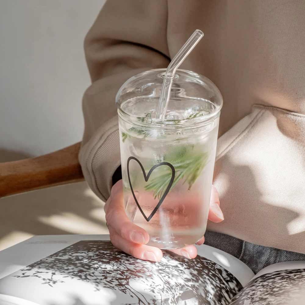 Eine Person hält einen durchsichtigen Becher mit Herzmotiv, Kräutern, Eis und einem Glasstrohhalm mit dem Eulenschnitt-Glasdeckel für Trinkglas und hat ein offenes Buch auf dem Schoß.