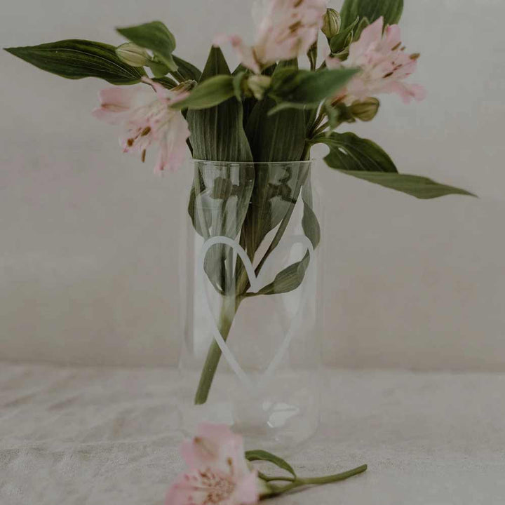 Das Eulenschnitt - Hohes Trinkglas Herz, ein klares Glas mit einem weißen Herz, enthält rosa Blumen und grüne Blätter; eine rosa Blume mit Stiel liegt vor dem Glas.