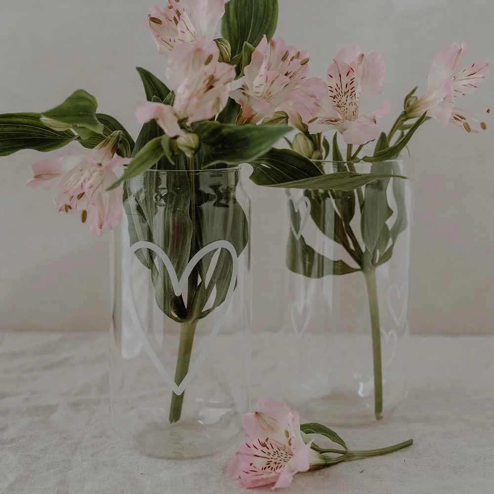 Zwei Eulenschnitt-Gläser – Hohes Trinkglas Herz, jeweils mit einem weißen Herzmuster, enthalten rosa Blumen und grüne Blätter; eine rosa Blume liegt auf der Oberfläche neben den Gläsern.