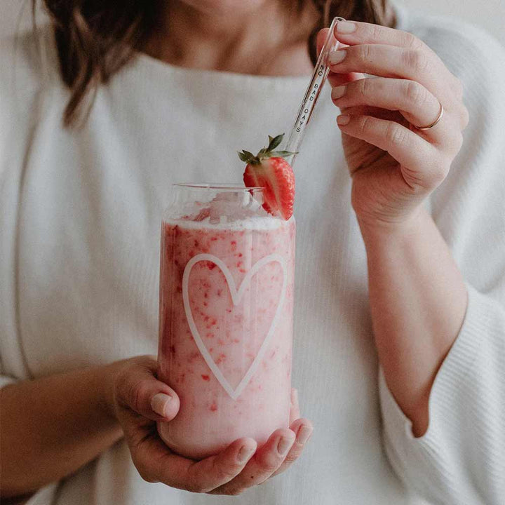 Jemand hält ein Eulenschnitt - Hohes Trinkglas Herz, gefüllt mit rosa Erdbeermilch, dekoriert mit einem Herzdesign, garniert mit Schlagsahne und einer Erdbeere, und rührt es mit einem durchsichtigen Strohhalm um.