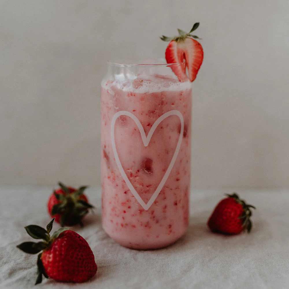 Ein Glas rosa Erdbeermilch mit Eis im Eulenschnitt – Hohes Trinkglas Herz, garniert mit einer Erdbeerscheibe und einer weißen Herzkontur, umgeben von ganzen Erdbeeren.
