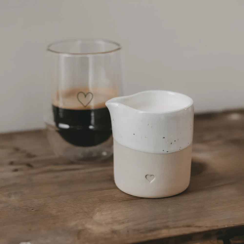 Ein Glas dunklen Kaffees steht neben dem Eulenschnitt - Kännchen Herz, einem Milchkännchen aus Keramik mit Herzmuster, auf einer Holzoberfläche.