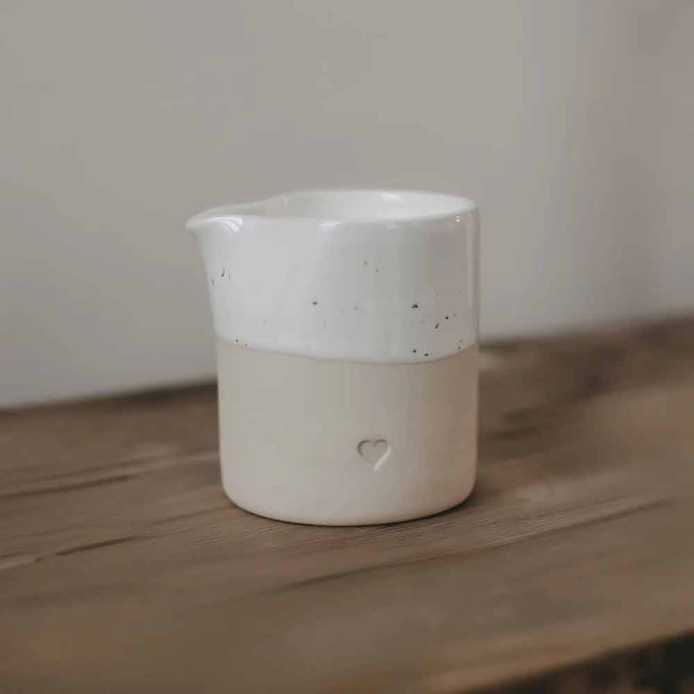 Das Eulenschnitt - Kännchen Herz ist ein kleiner Keramikkrug mit Ausguss, oben weiß und unten beige, mit einer Herzvertiefung auf einer Holzoberfläche.