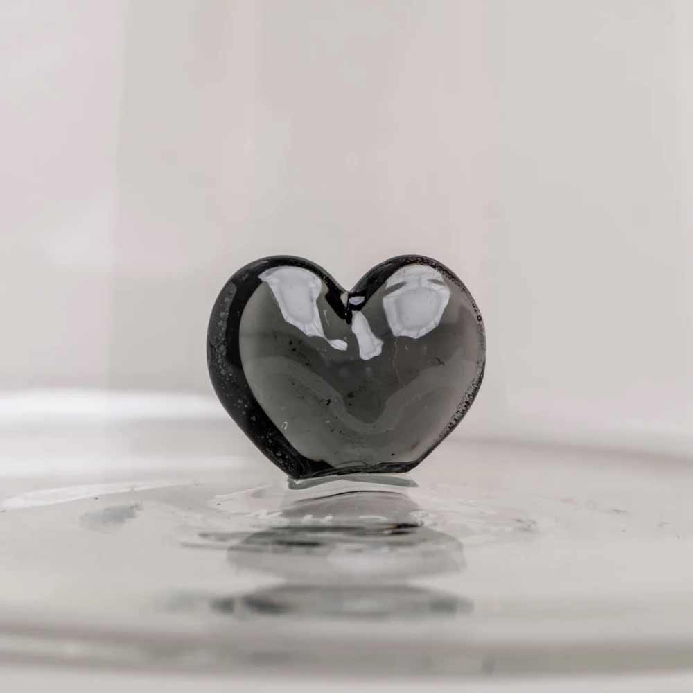 Die Eulenschnitt - Karaffe Herzfigur schwarz, eine glänzend schwarze Karaffe in Herzform, ruht elegant auf einer reflektierenden Oberfläche vor einem schlichten, hellen Hintergrund.