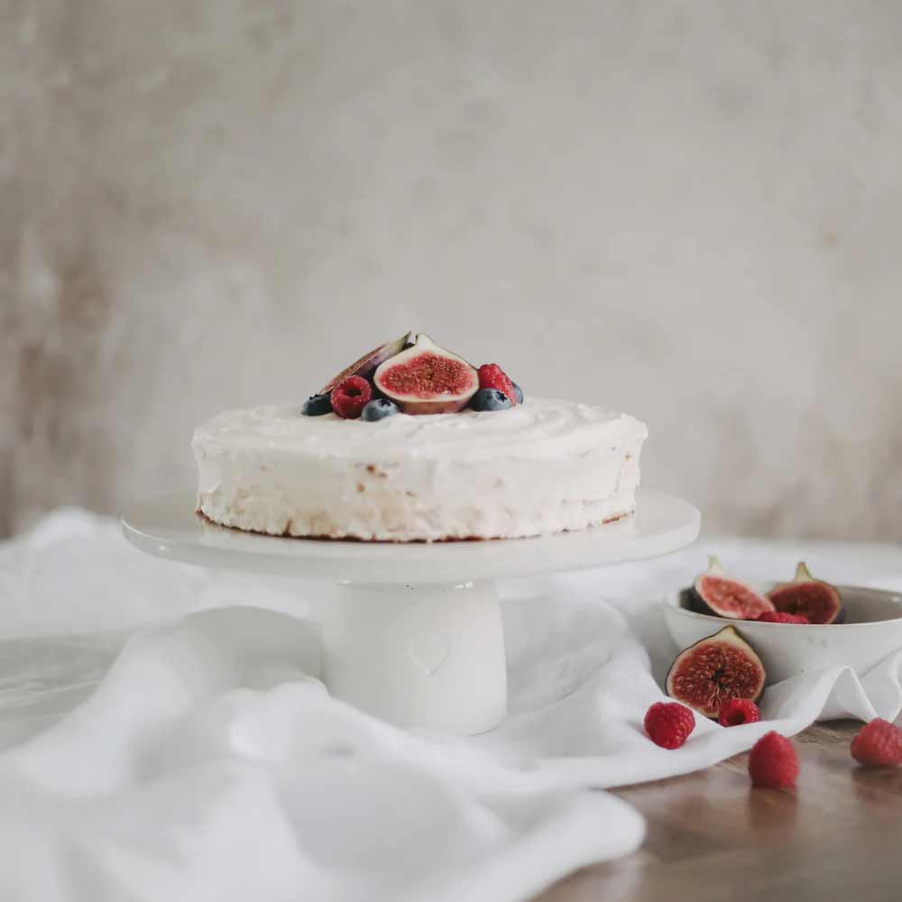 Auf dem Eulenschnitt - Kuchenständer Herz 10 cm steht ein runder Kuchen mit weißem Zuckerguss und Feigen, Himbeeren und Blaubeeren, daneben steht in einer kleinen Schüssel zusätzliches Obst.