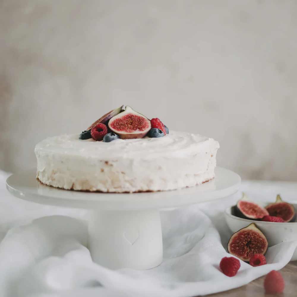Auf dem Eulenschnitt - Kuchenständer Herz 10 cm steht ein runder, weiß glasierter Kuchen mit Feigen, Himbeeren und Blaubeeren, daneben liegen noch weitere Feigen und Beeren.