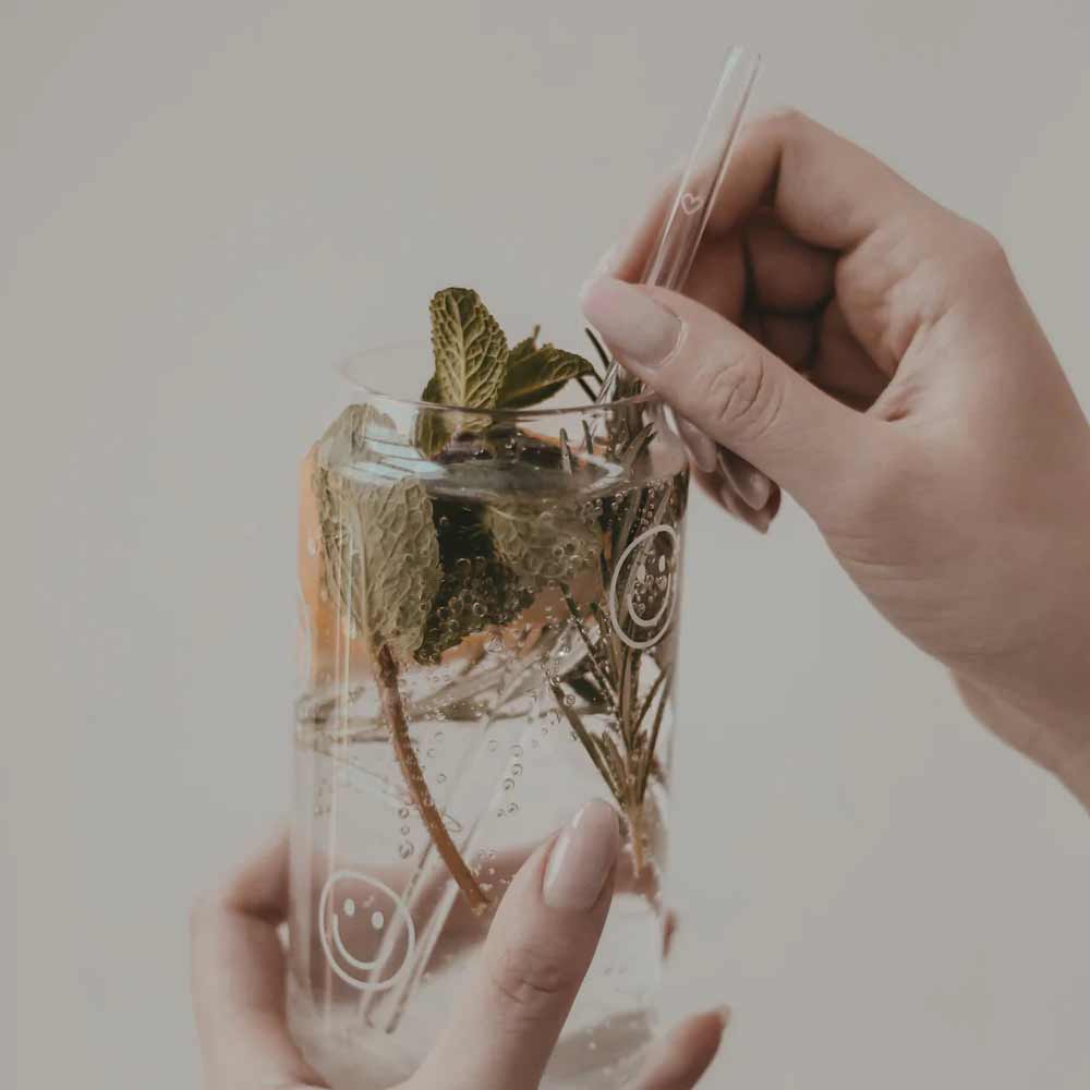 Eine Person hält ein klares Getränk mit Kräutern in einem mit einem Smiley bedruckten Glas und nippt durch einen Eulenschnitt-Glashalm aus dem 4er-Set „Strohhalme Herz aus Glas 20cm“.