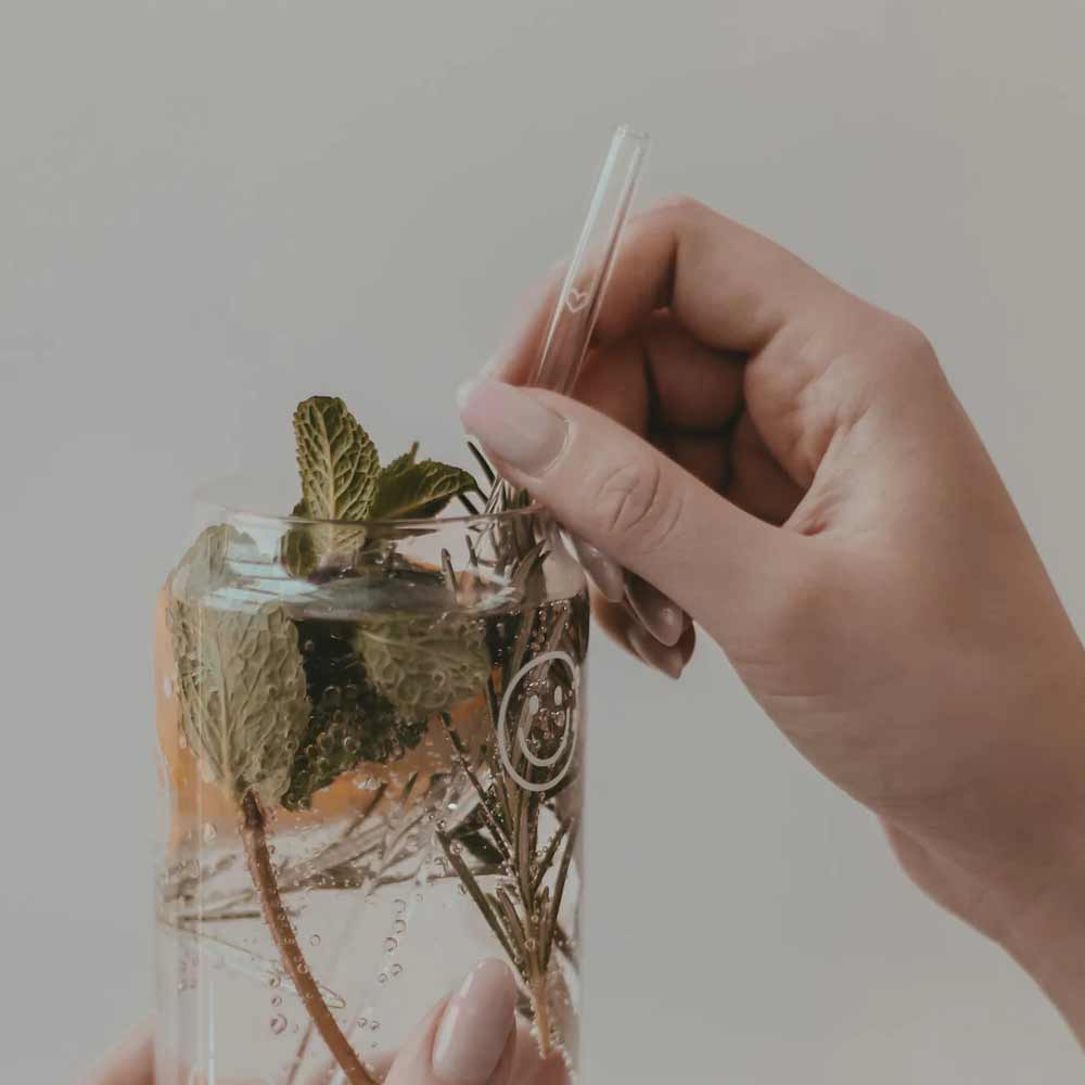 Eine Hand hält ein Glas mit einem klaren Strohhalm Eulenschnitt - Strohhalme Herz aus Glas 20cm (4er Set), gefüllt mit Sprudelwasser, Eis, Minzblättern und Rosmarinzweigen.