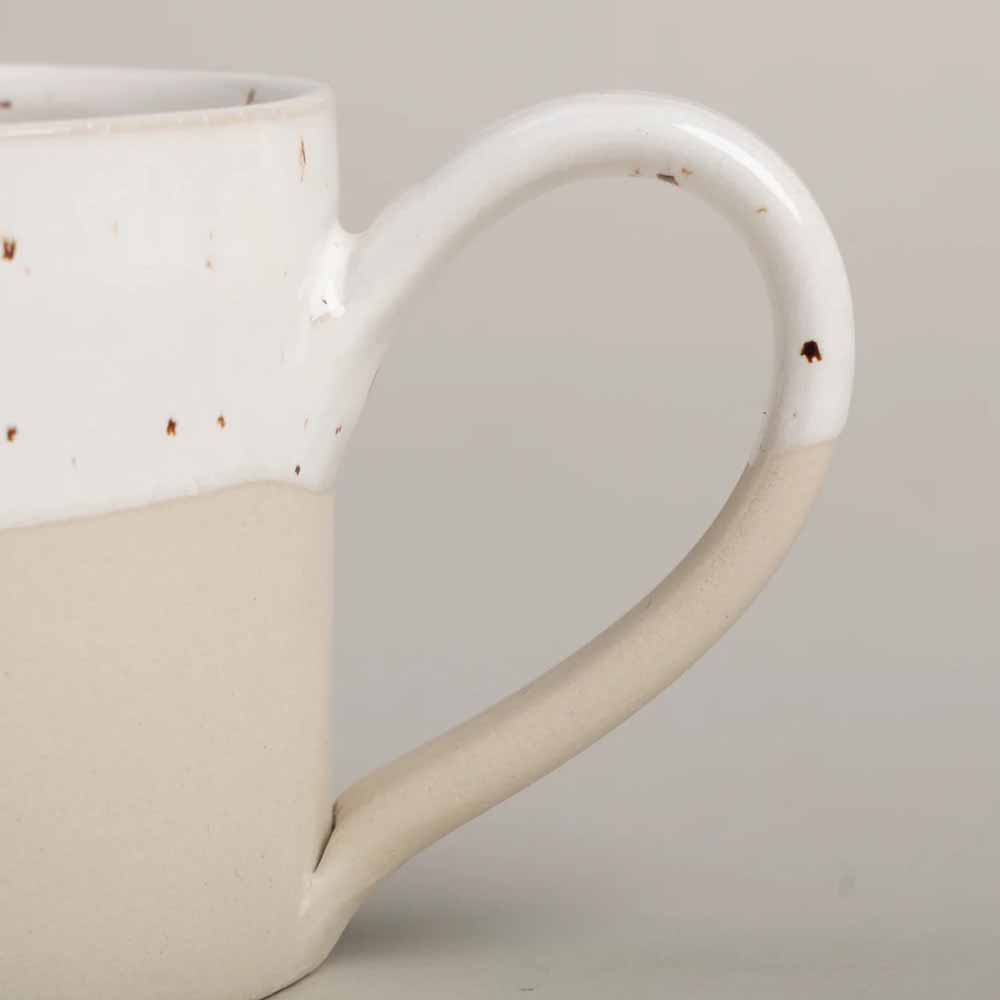 Eine zweifarbige Keramiktasse mit einem weißen Oberteil und einem beigen Unterteil. Der Henkel ist ebenfalls zweifarbig, passend zu den Farben der Tasse. Die Oberfläche weist einige dunklere Flecken auf, die Eulenschnitt - Tasse Herz genannt werden.