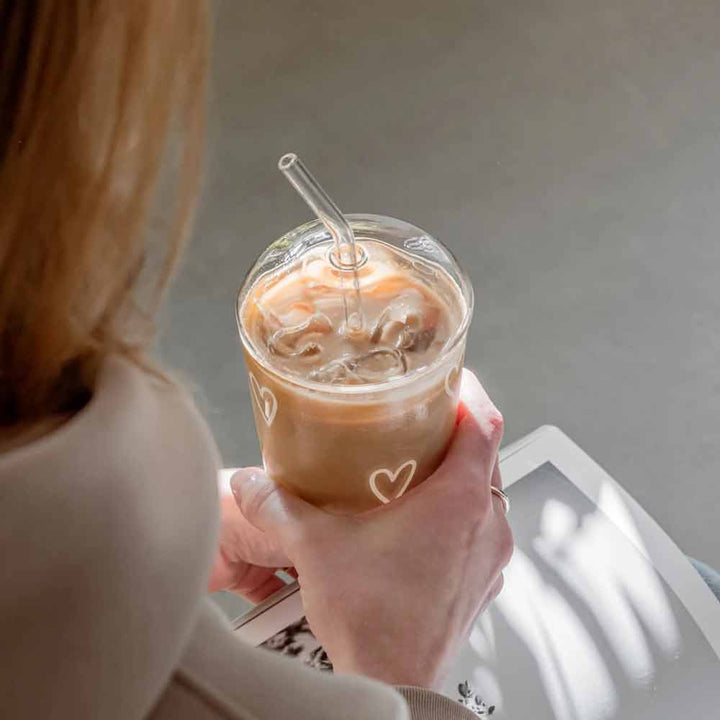 Eine Person hält ein Eulenschnitt-Trinkglas Herzen weiß, gefüllt mit Eiskaffee und einem durchsichtigen Strohhalm, von oben und hinten betrachtet, während das Sonnenlicht Schatten auf das Tablett darunter wirft.