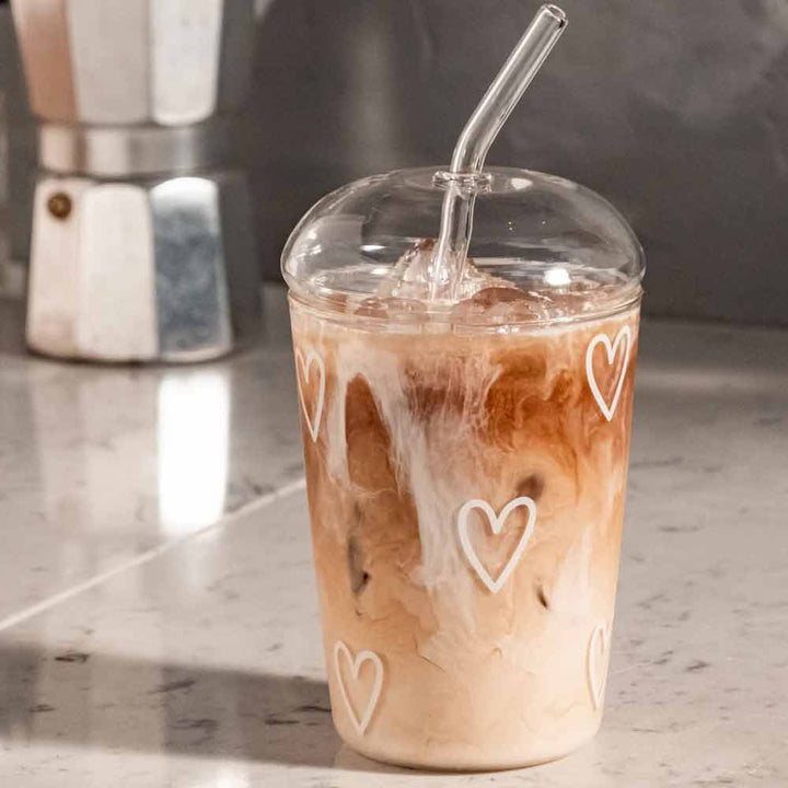 Ein Eiskaffee mit Milch wird im Eulenschnitt - Trinkglas Herzen weiß gewirbelt, einer durchsichtigen Tasse, die mit weißen Herzen und einem Glasstrohhalm verziert ist und auf einer Marmortheke steht.