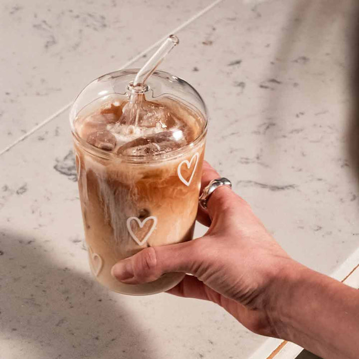 Eine Hand hält ein Eulenschnitt-Trinkglas Herzen weiß mit Eiskaffee und Milch, mit weißem Herzmotiv, einem durchsichtigen Deckel und einem Glasstrohhalm, platziert auf einer Marmoroberfläche.