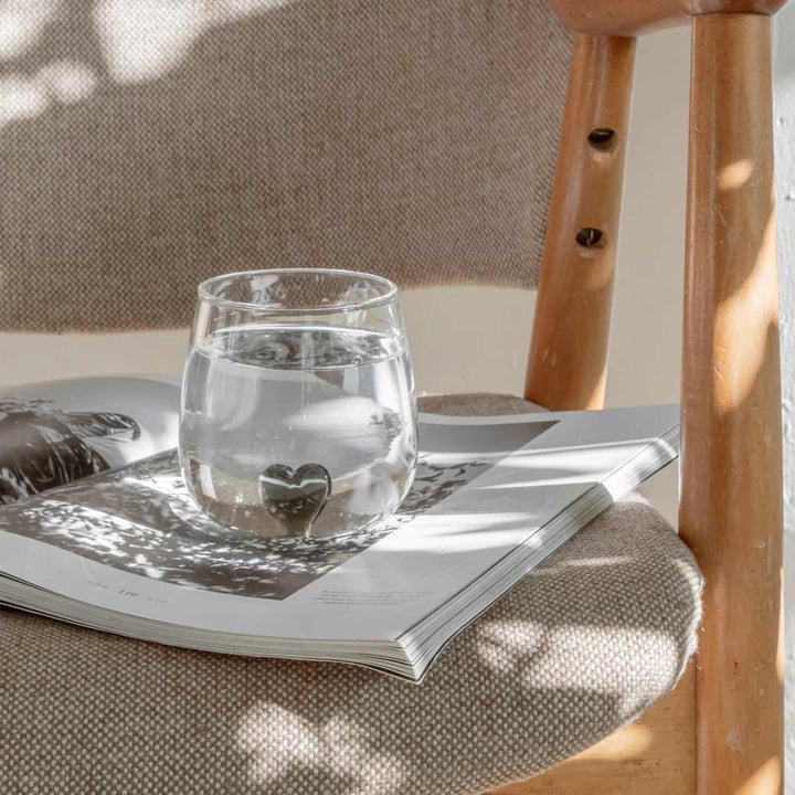 Eine schwarze Herzfigur aus dem Eulenschnitt-Trinkglas liegt auf einer offenen Zeitschrift auf einem gepolsterten Holzstuhl, wobei sanftes natürliches Licht sanfte Schatten erzeugt.