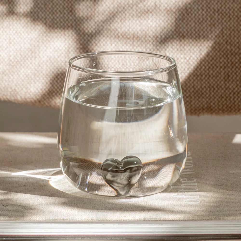Eine Eulenschnitt - Trinkglas Herzfigur schwarz mit herzförmigem Sockel steht auf einem geschlossenen Buch, Sonnenlicht und Schatten heben das klare Glasdesign hervor.