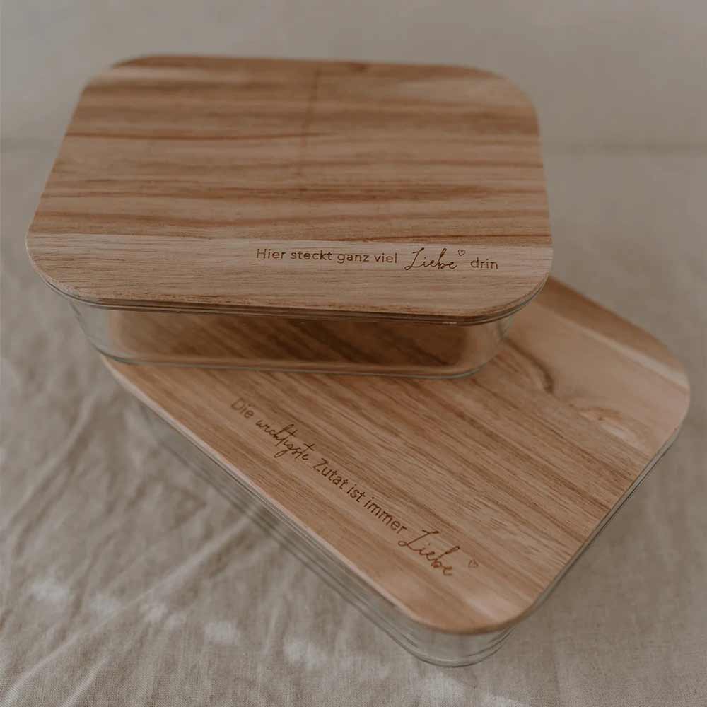 Zwei Eulenschnitt-Vorratsbehälter aus Glas mit graviertem Holzdeckel und Spruch, jeweils mit dem Wort „Liebe“, sind übereinandergestapelt auf neutralem Stoff zu sehen.