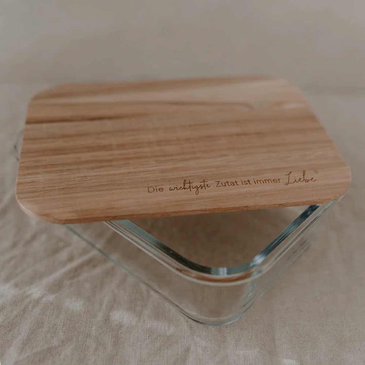 Der Eulenschnitt - Vorratsbehälter Glas mit Holzdeckel und Spruch verfügt über einen Glasbehälter mit Holzdeckel, in den auf Deutsch „Die wichtigste Zutat ist immer Liebe“ eingraviert ist, und ist auf einer beigen Stoffoberfläche abgebildet.
