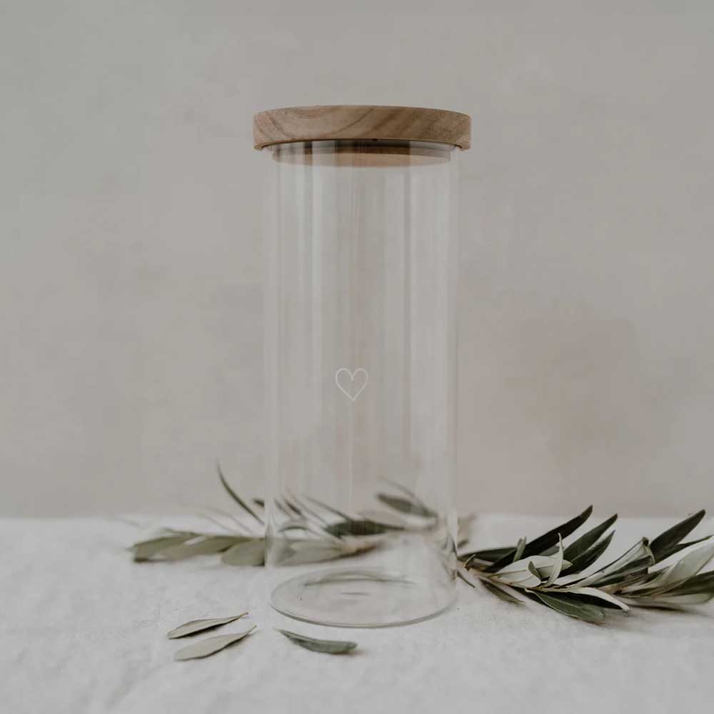 Das Eulenschnitt - Vorratsglas Herz 25cm, ein klares Glasgefäß mit Holzdeckel, ruht auf einer weißen Fläche inmitten von Olivenzweigen.