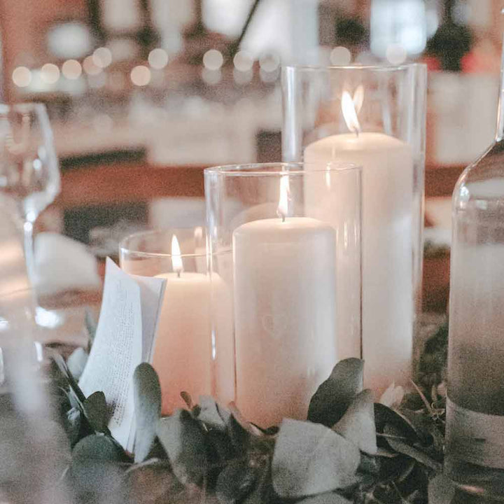 Drei weiße Stumpenkerzen brennen in 25 cm großen Vasen aus Vorratsglas „Herz“ von Eulenschnitt, umgeben von Grünpflanzen und gefaltetem Papier, mit verschwommenem Glasgeschirr im Hintergrund.