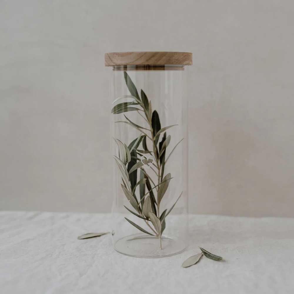 Das Eulenschnitt - Vorratsglas Herz 25cm mit Holzdeckel enthält Olivenzweige und steht auf einem weißen Tuch, neben dem Glas liegen mehrere Blätter verstreut.