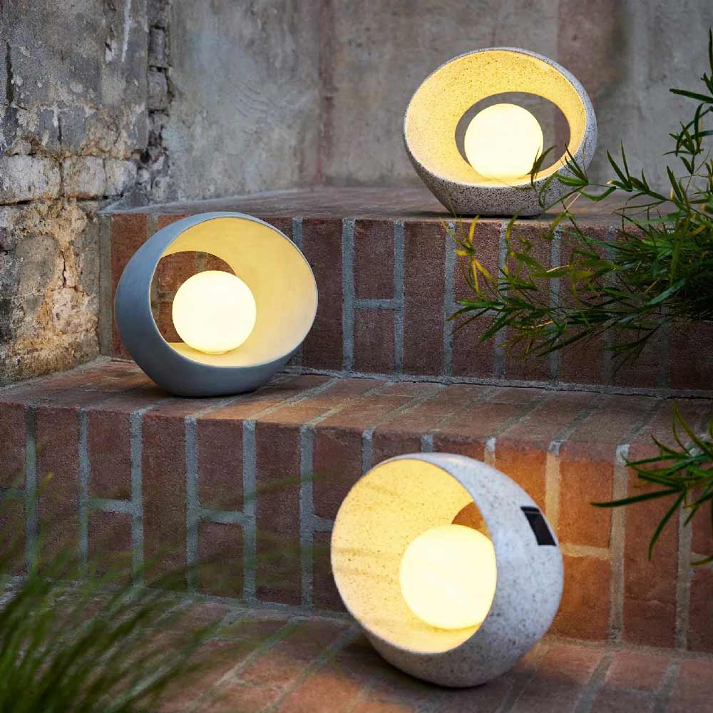 Drei Gilde - Laterne Solar Ovalo Außenlampen mit runden Steingehäusen stehen auf Ziegelstufen neben einer verwitterten Mauer und grünen Pflanzen.